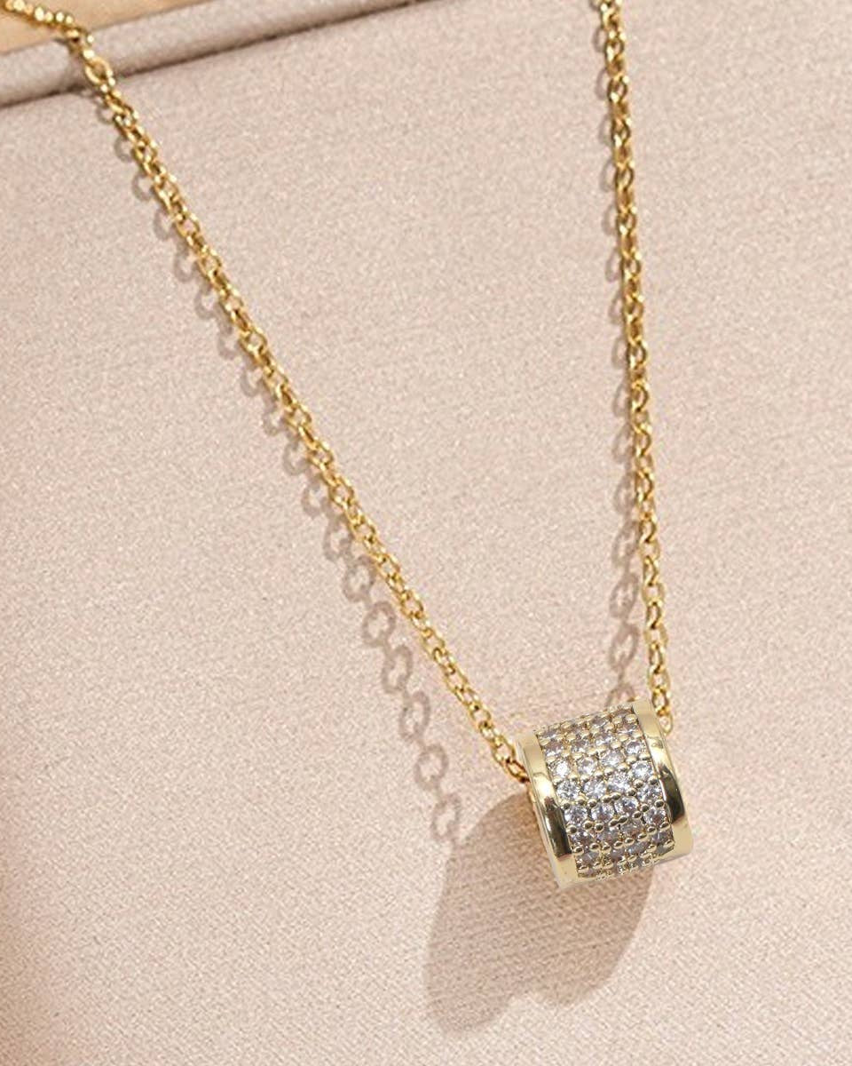 A gold chain necklace with a cylindrical pendant encrusted with small clear stones, displayed on a light beige jewelry box.