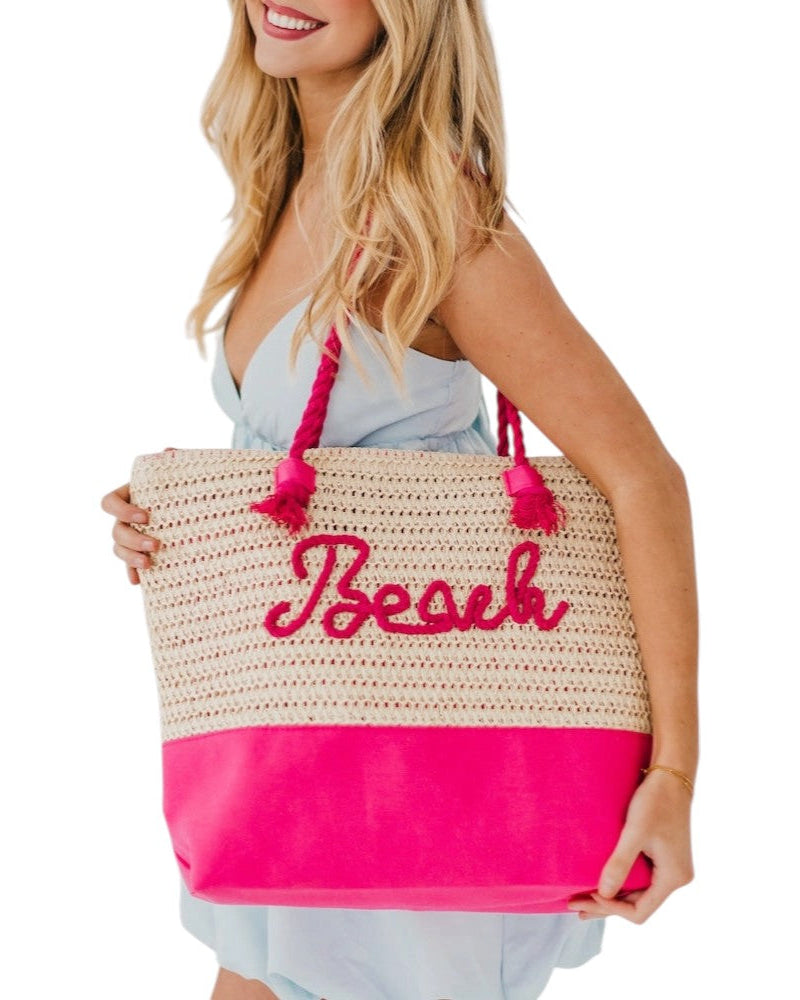 Woman holding a straw beach bag with pink accents and 'Beach' text on a white background.