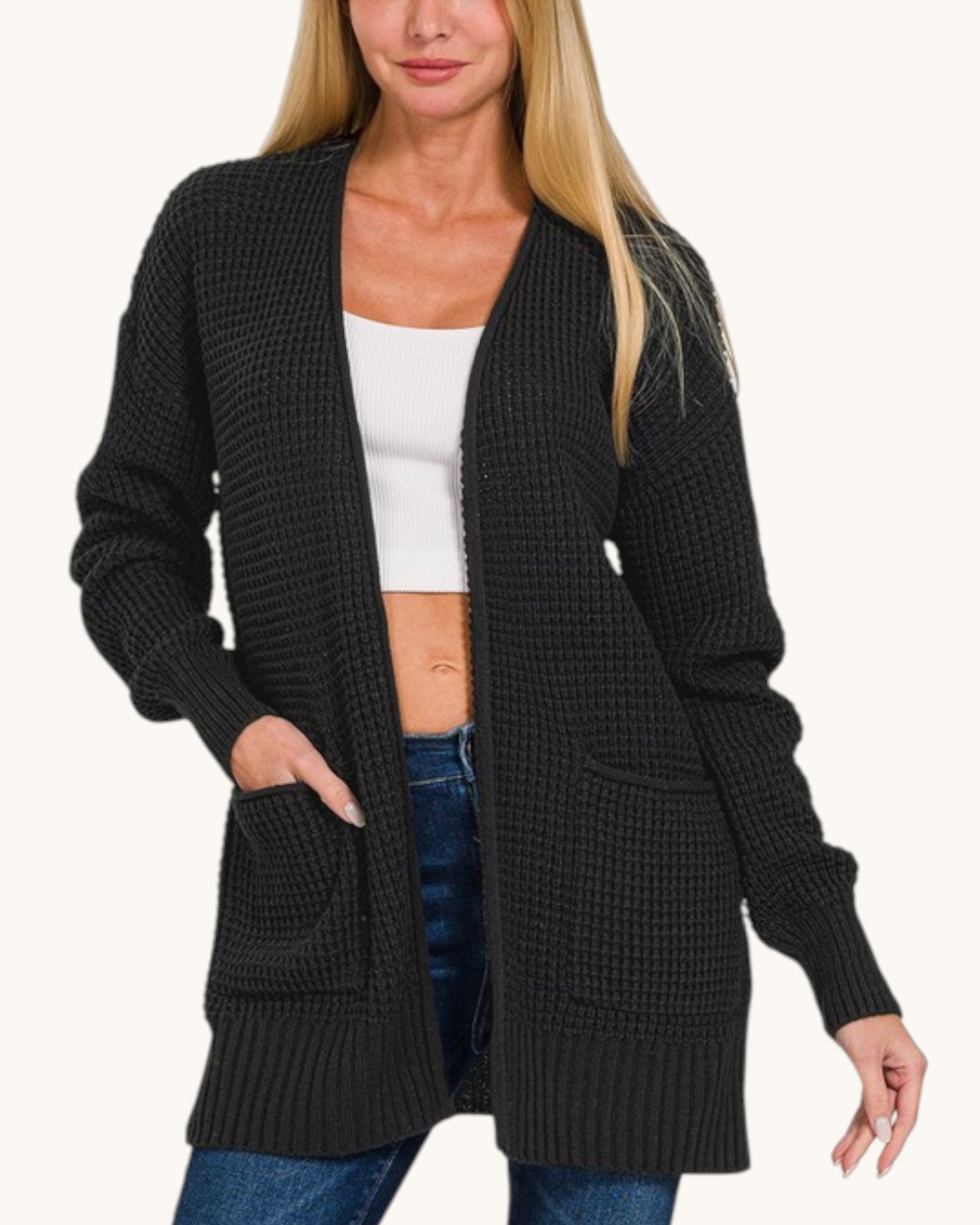 A woman wearing a black waffle-knit open-front cardigan with long sleeves and front patch pockets, styled over a white crop top and blue jeans against a plain light background.