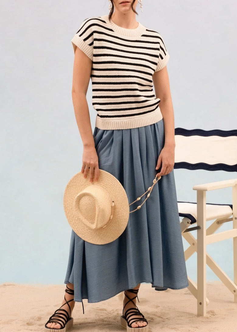 Woman wearing a striped short sleeve cream and black knit sweater and blue skirt standing on sand with a chair in the background.