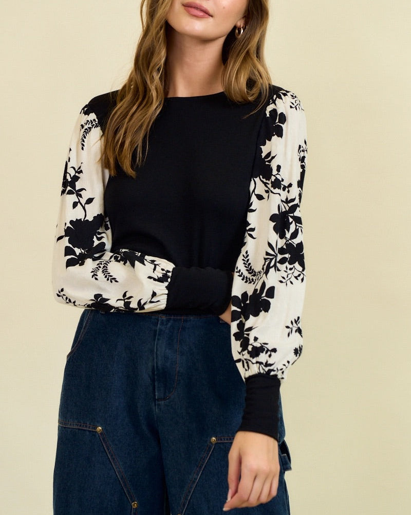Woman wearing a black and white floral top with blue jeans on a beige background.