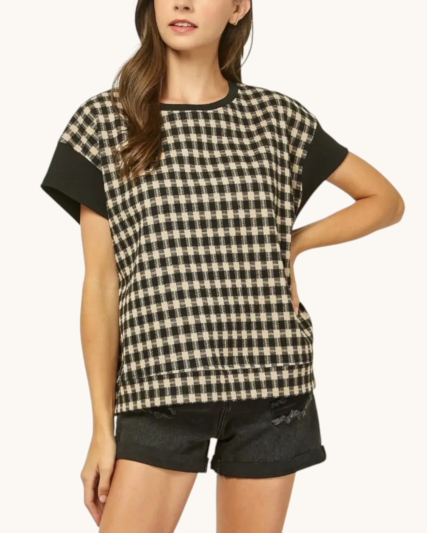 A woman wearing a short-sleeve black and beige gingham patterned top with solid black sleeve panels and black denim shorts, photographed against a plain light background.