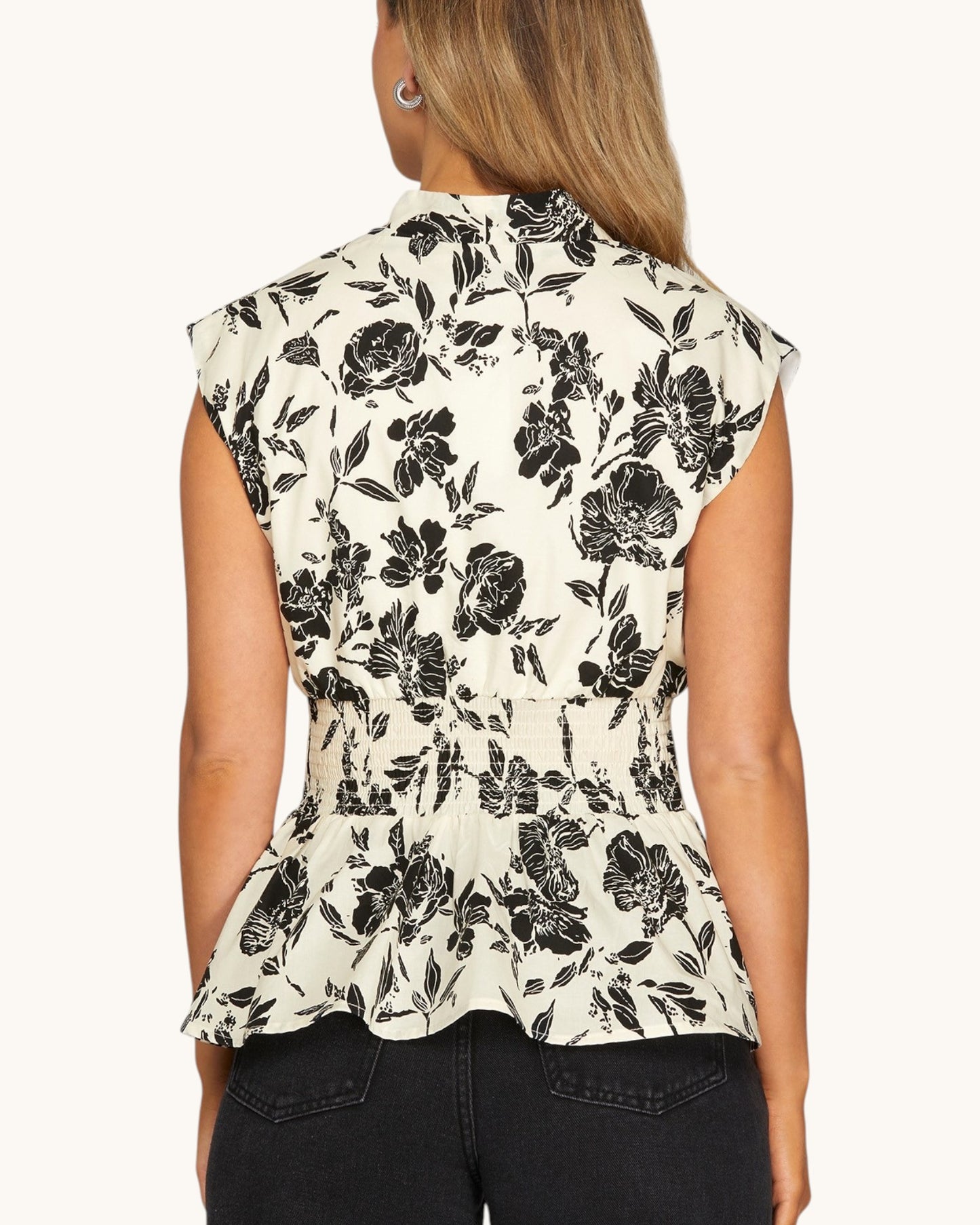 A woman shown from the back wearing a sleeveless peplum-style blouse with a black floral print on a warm off-white background, paired with dark jeans against a plain light background.