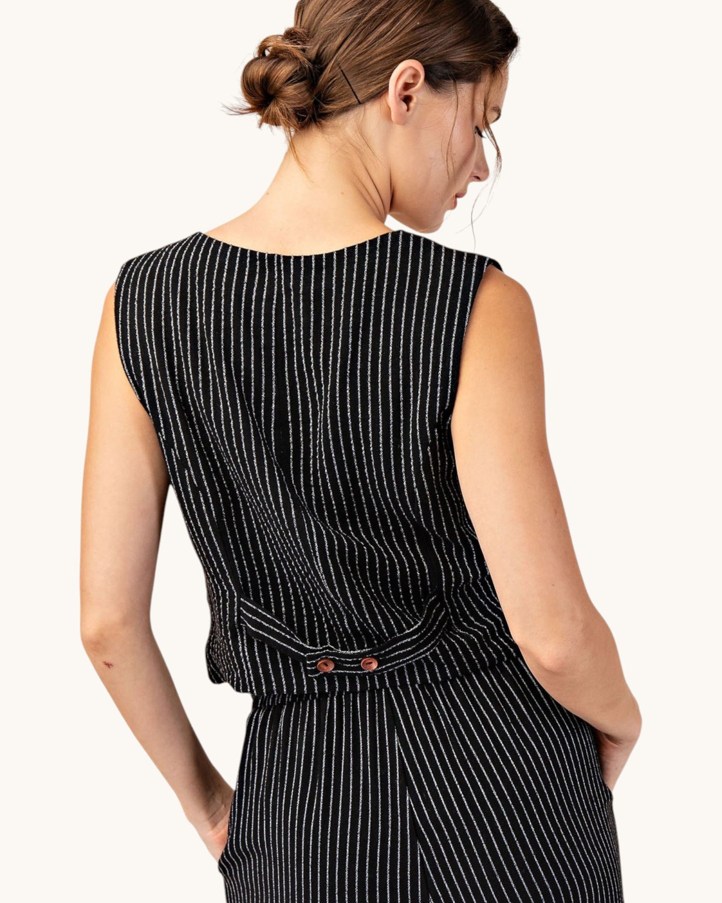 Back view of a woman wearing a cropped, fitted black pinstripe sleeveless button up vest and matching pants.