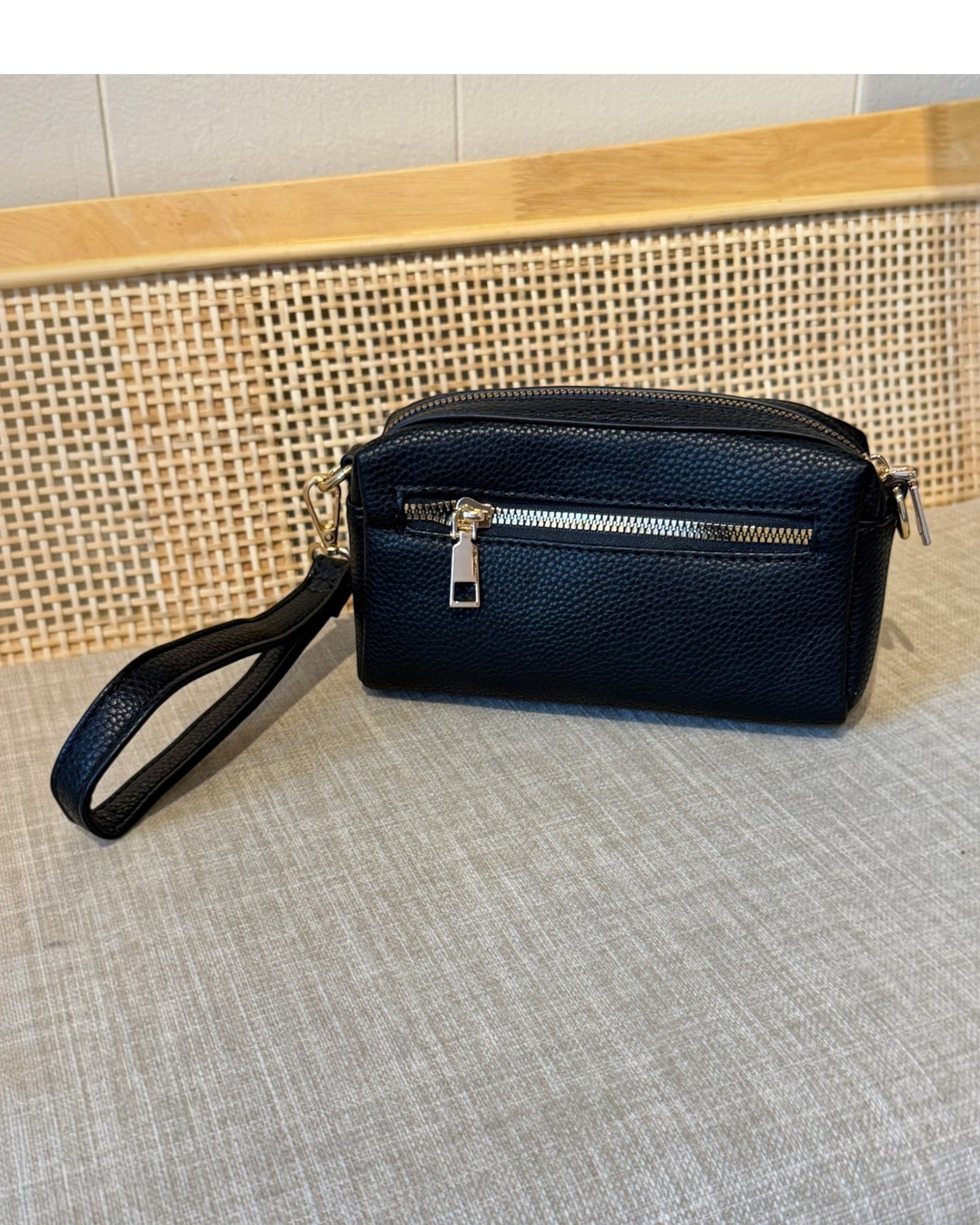 A black pebbled leather wristlet bag with a wrist strap and silver zipper, resting on a textured gray fabric surface in front of a woven cane panel.