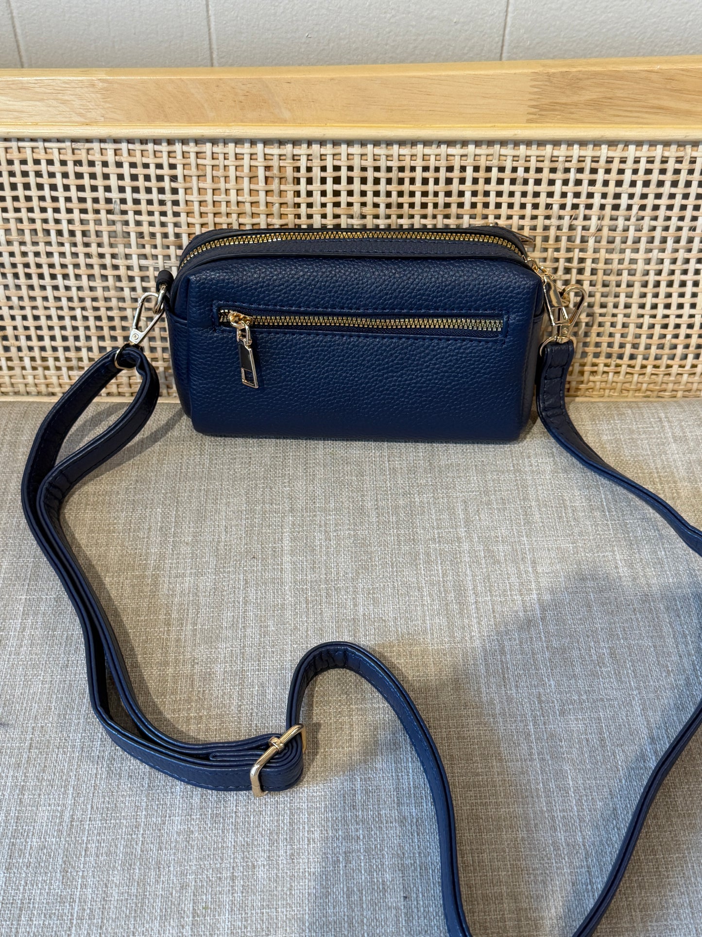 A navy blue pebbled leather crossbody bag with gold-tone hardware, featuring a top zip closure and a detachable shoulder strap, displayed on a textured light gray fabric surface in front of a woven cane panel.