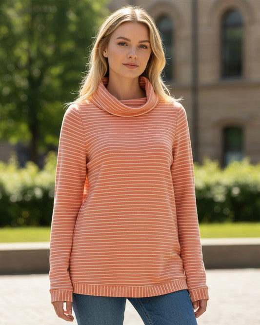 Woman wearing a salmon-color striped sweater tunic standing outdoors with greenery and a building in the background.