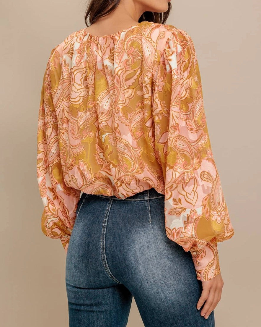 Woman wearing a camel, pink and ivory floral paisley print blouse and jeans against a beige background.