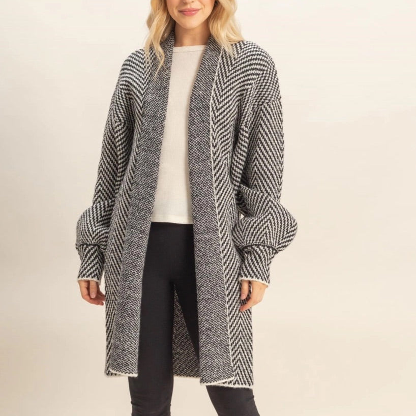 Woman wearing a long, B&W herringbone patterned cardigan over a white top and black pants on a beige background.