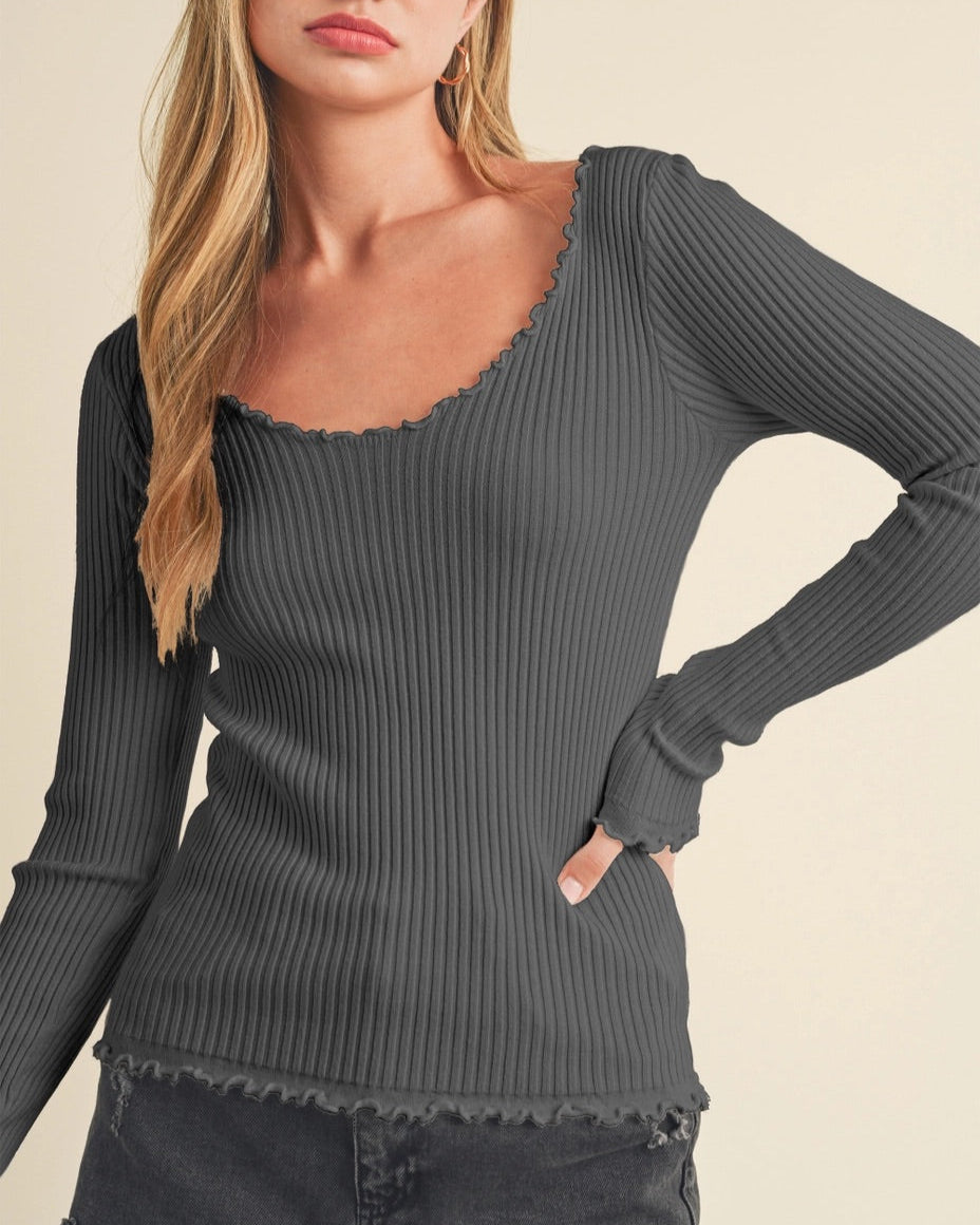 Woman wearing a slim fit dark gray ribbed long sleeve shirt with lettuce trim against a beige background.