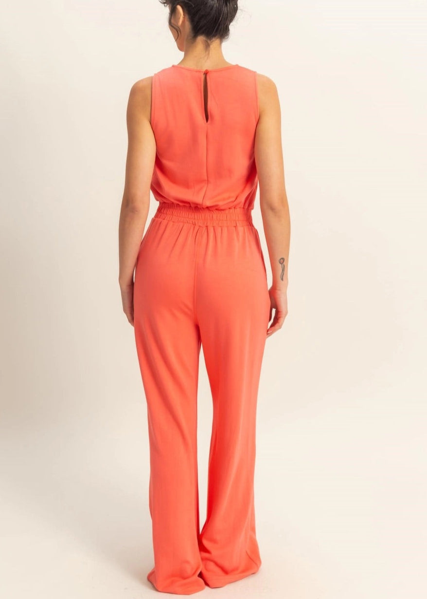Woman wearing Sunset Hour Sleeveless Jumpsuit in Coral with cinched waist and high scoop neckline.