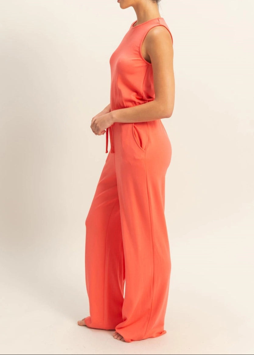 Woman wearing Sunset Hour Sleeveless Jumpsuit in Coral with cinched waist and high scoop neckline.
