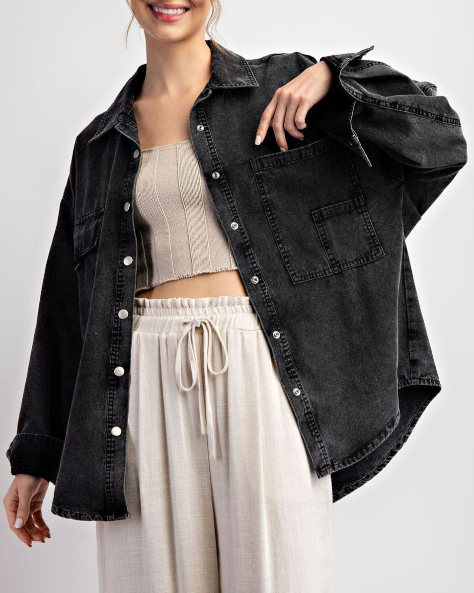 Woman wearing a black denim jacket / shacket over a beige top and white pants on a light gray background.