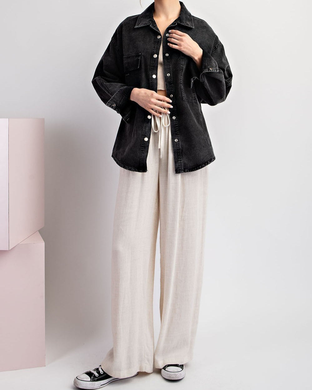 Woman wearing a black denim jacket / shacket and white pants on a light gray background.