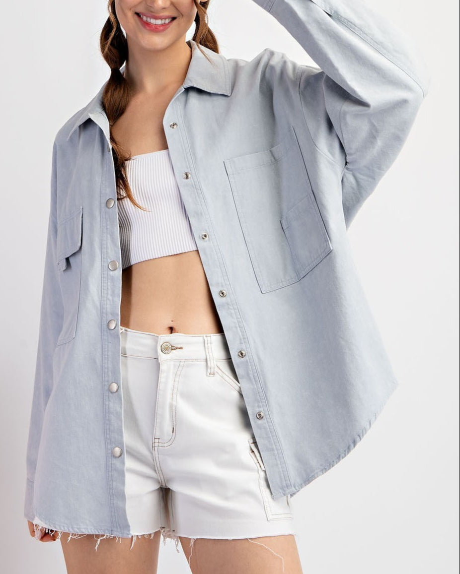 Woman wearing a light blue oversized denim shirt jacket /shacket and white shorts on a plain background.