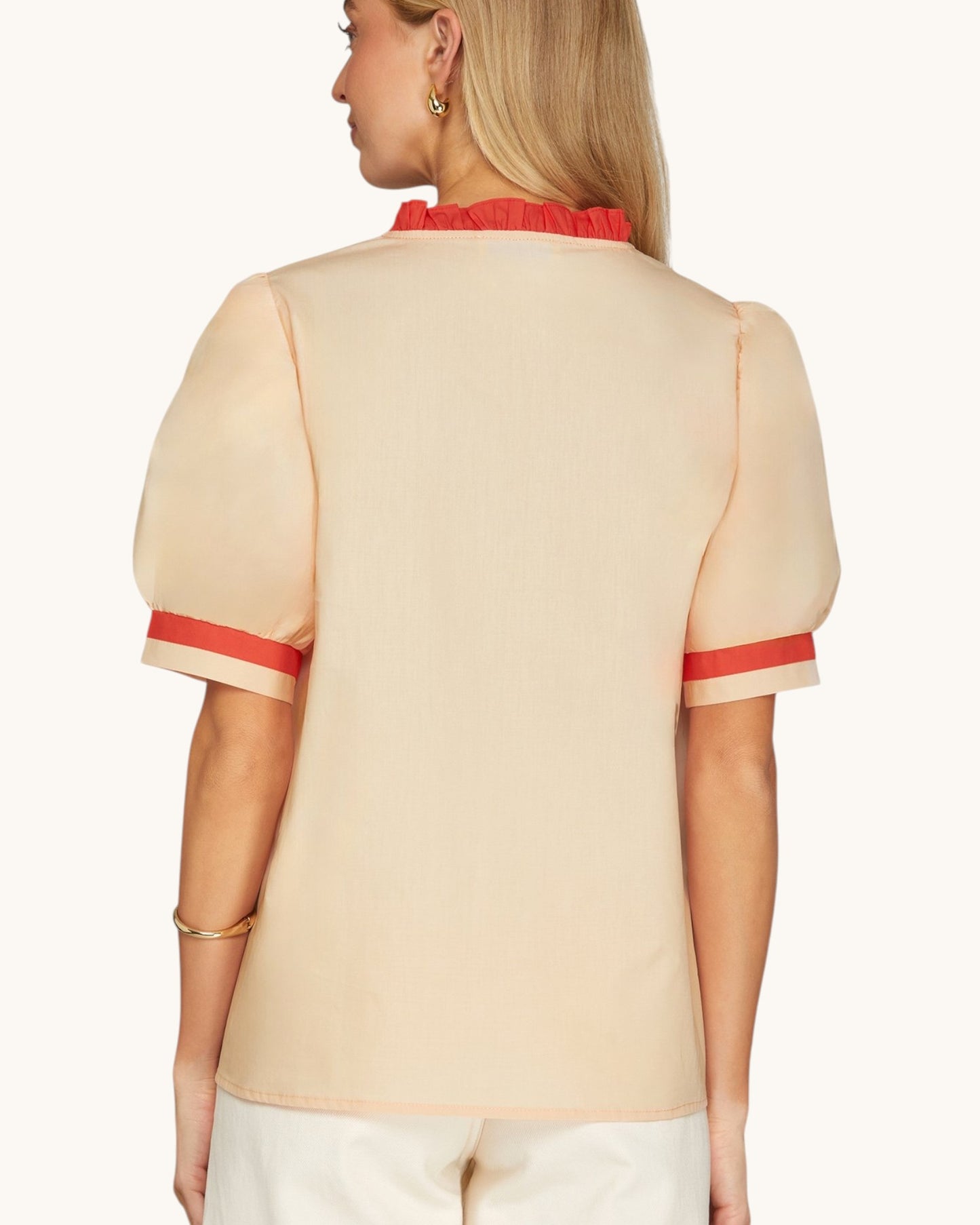 A woman shown from the back wearing a light beige short-sleeve blouse with a red ruffled collar and red stripe accents at the sleeve cuffs, paired with white pants against a light beige studio background.