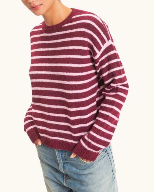 A woman wearing a burgundy and light pink striped knit sweater with light blue jeans against a plain off-white background.