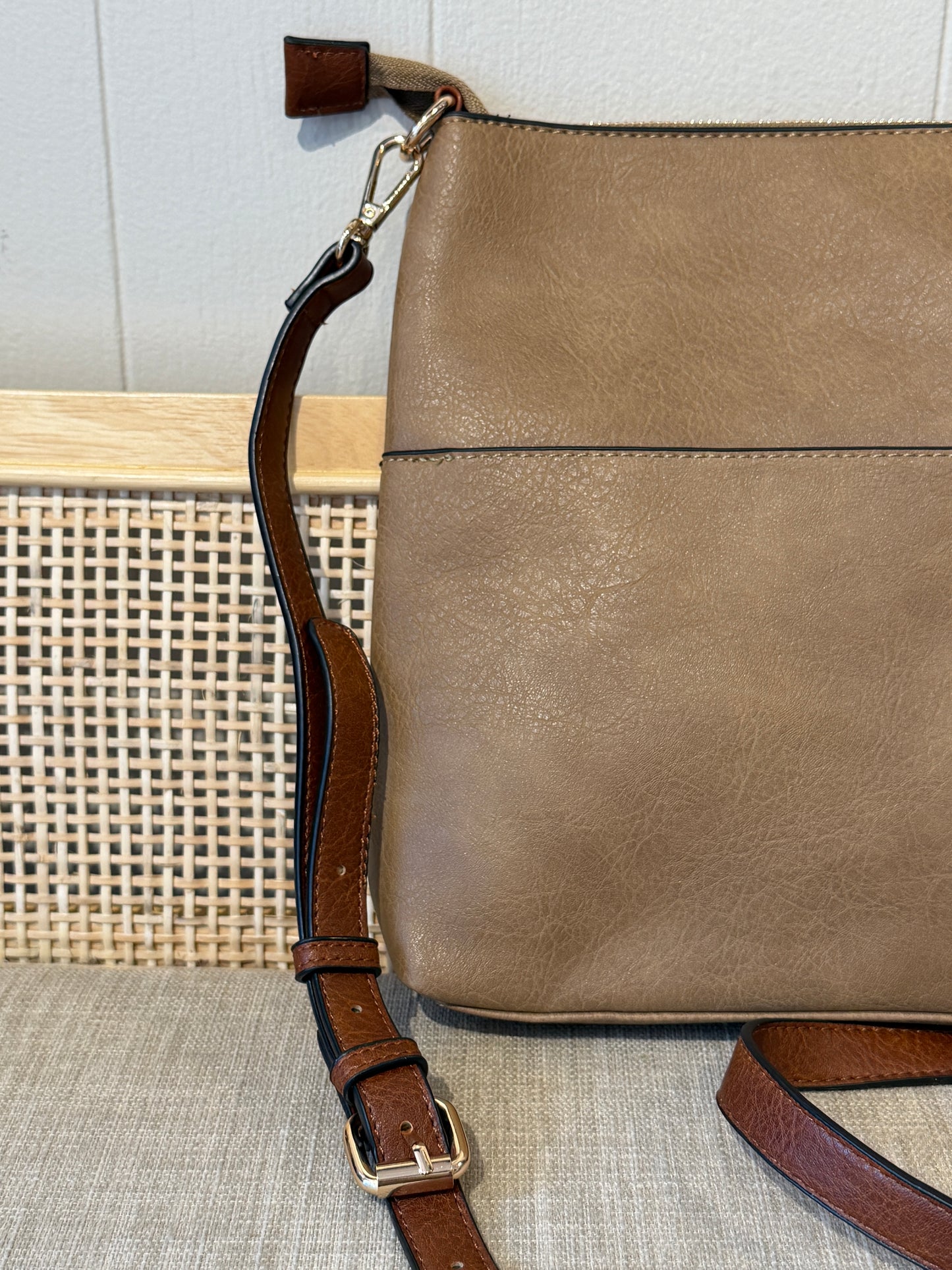 A taupe/mocha faux-leather shoulder bag with a brown leather strap and gold-tone hardware, shown resting on a woven cane chair against a light gray paneled wall.