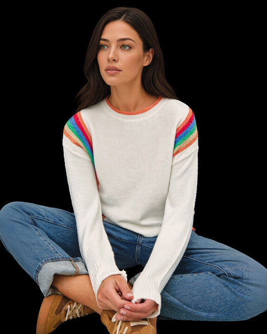 Seated model wearing ivory Rainbow Prism crewneck sweater with rainbow shoulder stripes and coral neckline trim.
