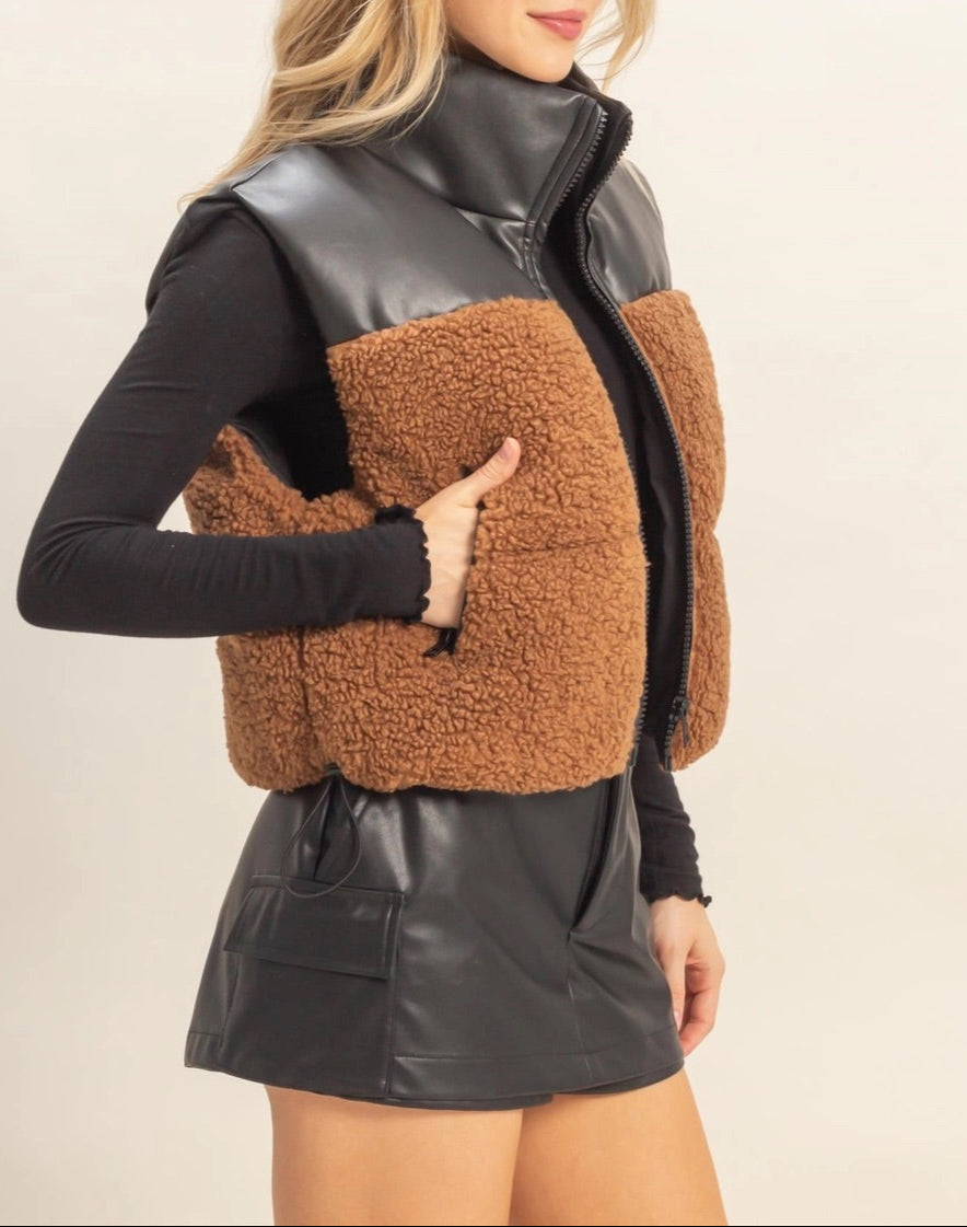 Woman wearing a brown sherpa and black faux leather vest with a plain background.