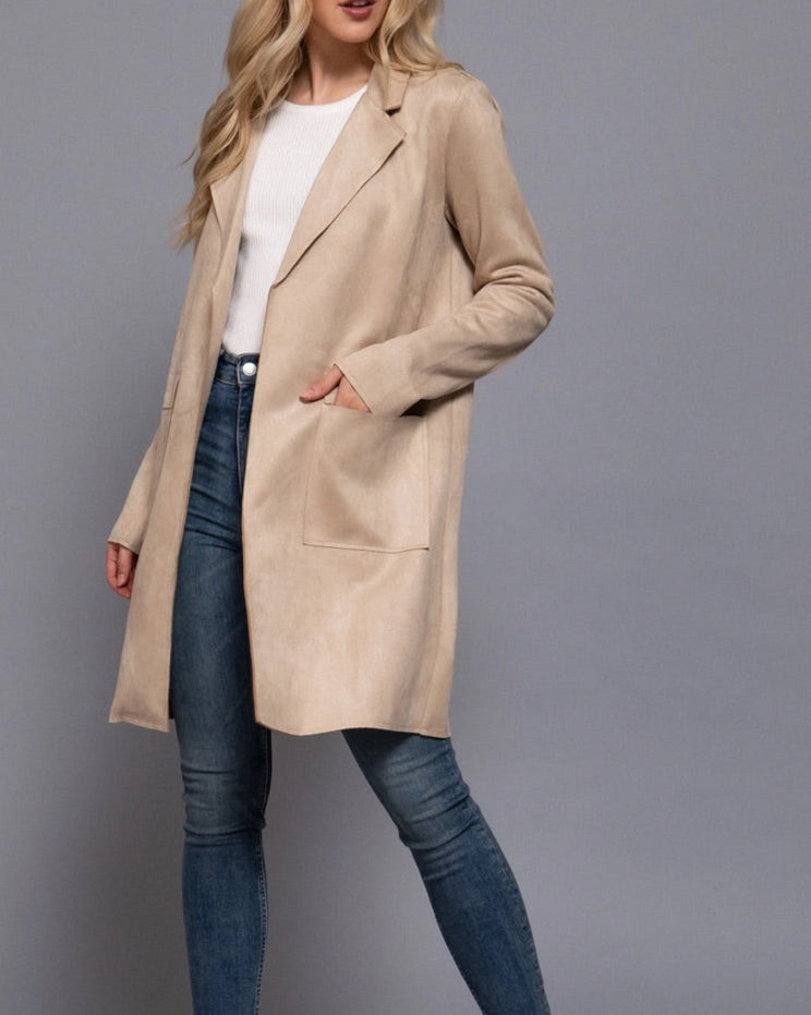 Woman wearing a beige coat, white shirt, blue jeans, and black boots on a gray background.