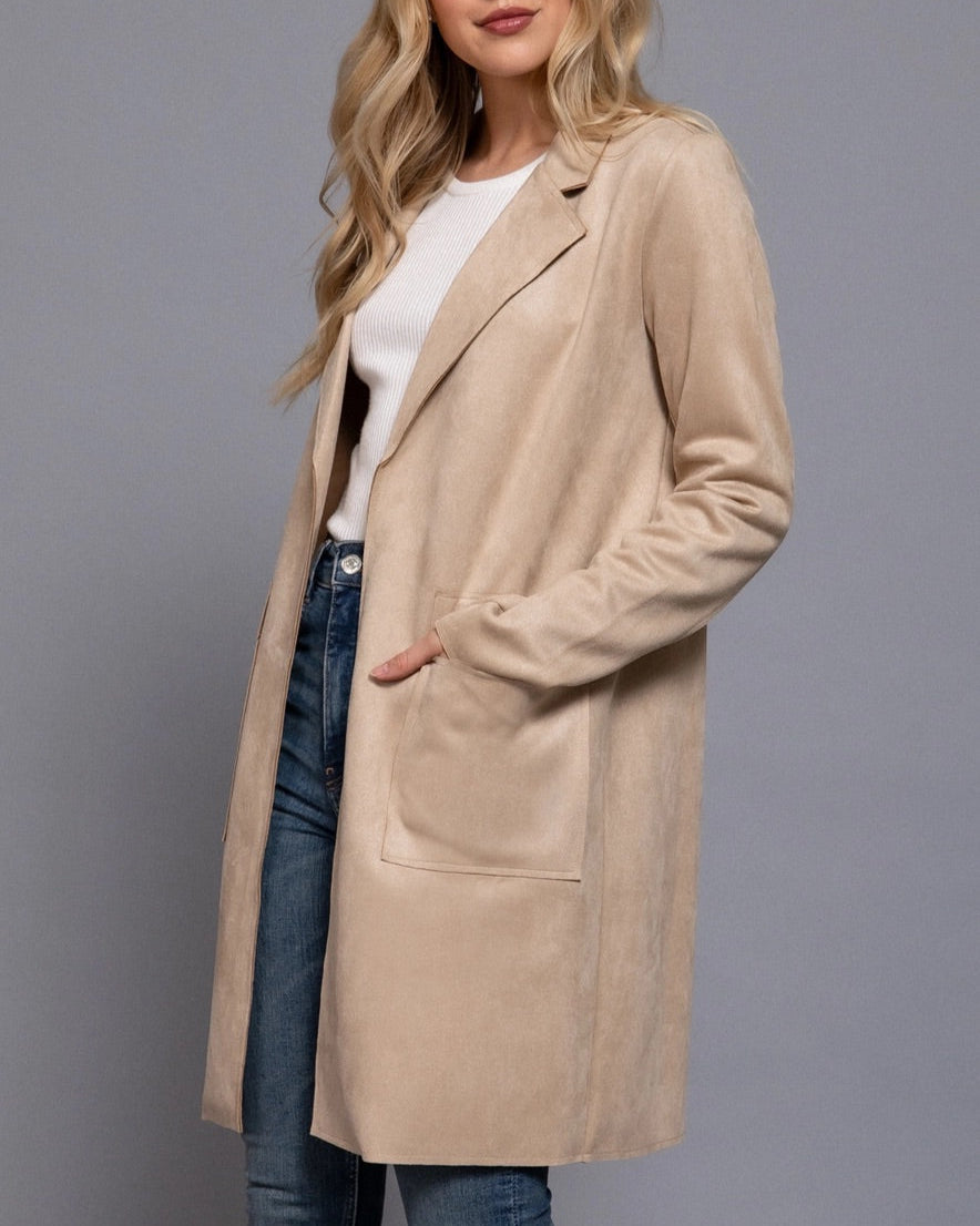 Woman wearing a beige coat over a white shirt and blue jeans on a gray background.