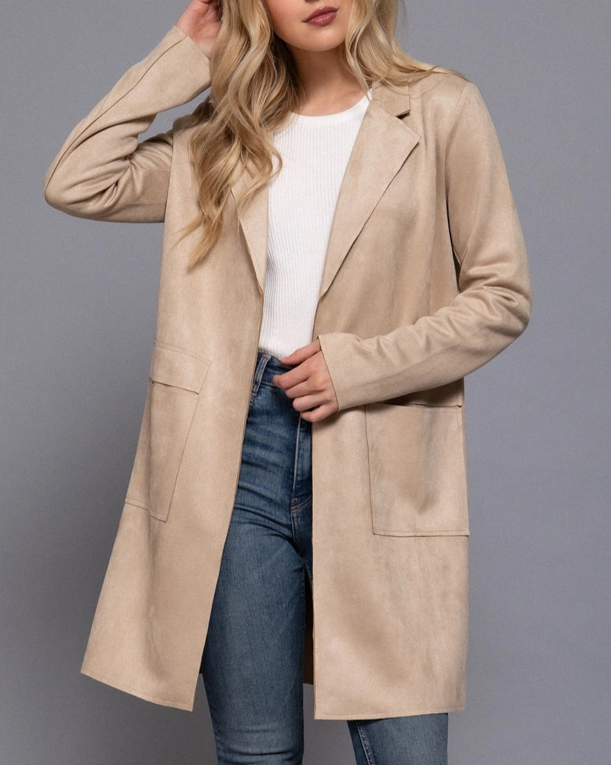 Woman wearing a sand faux suede coat over a white shirt and blue jeans on a gray background.
