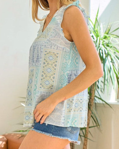 Woman wearing a green, blue and ivory geometric patterned sleeveless top and denim shorts.