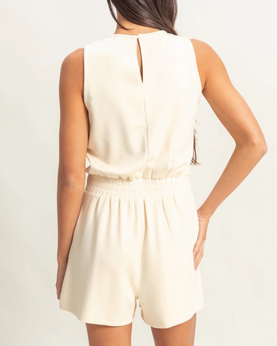 Woman wearing sleeveless cream lounge romper with drawstring shorts and keyhole back detail.
