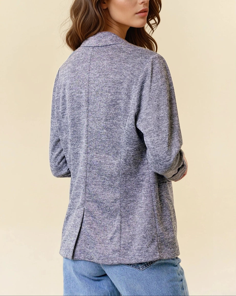 Woman wearing a charcoal blazer over a white top and blue jeans on a beige background.