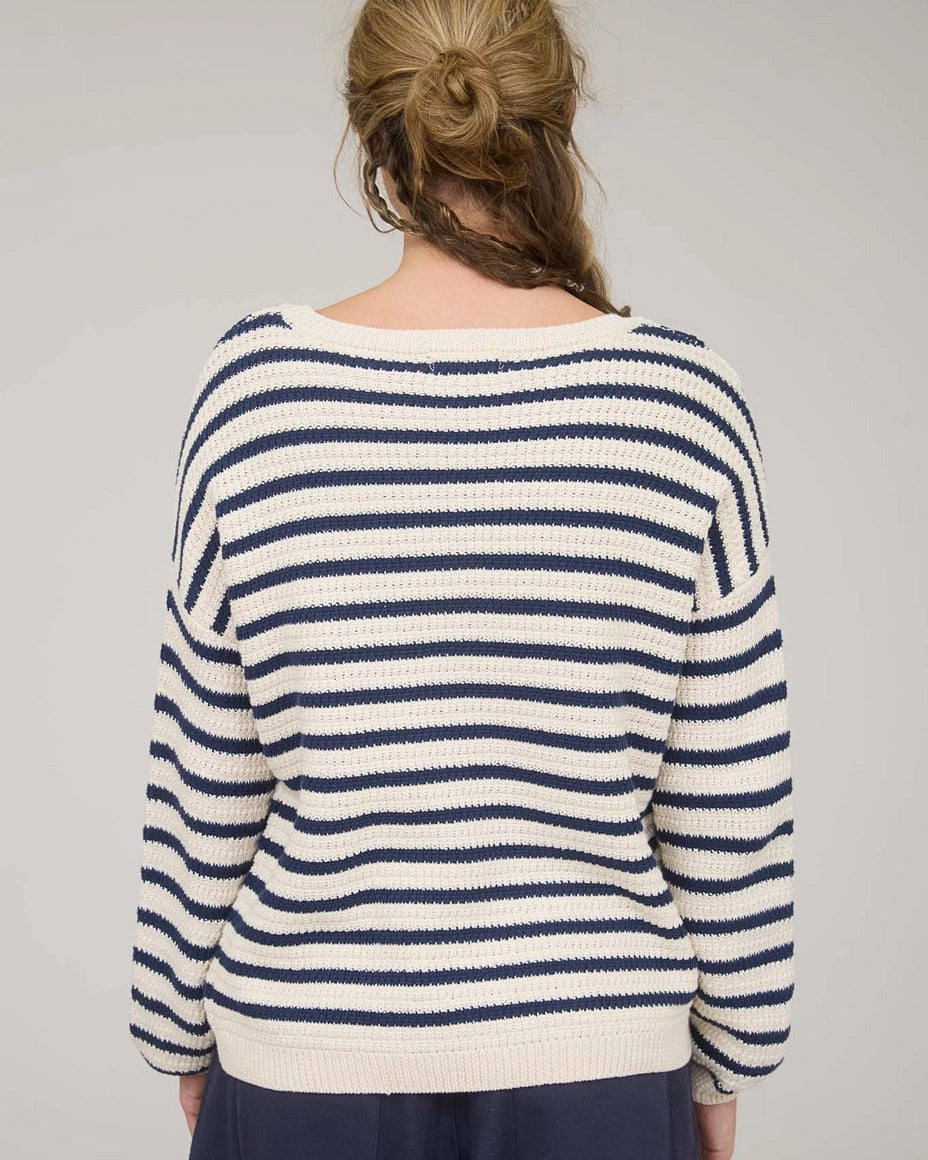 Rear view of a woman wearing a navy and white striped long sleeve sweater with navy pants against a plain background.