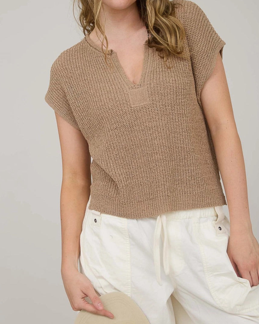 Woman wearing a tan knitted top and white pants against a plain background.