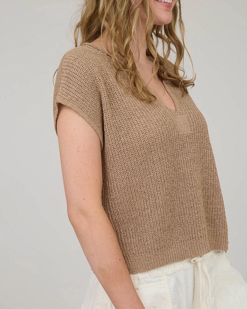 Woman wearing a tan knitted split V top and white pants against a plain background
