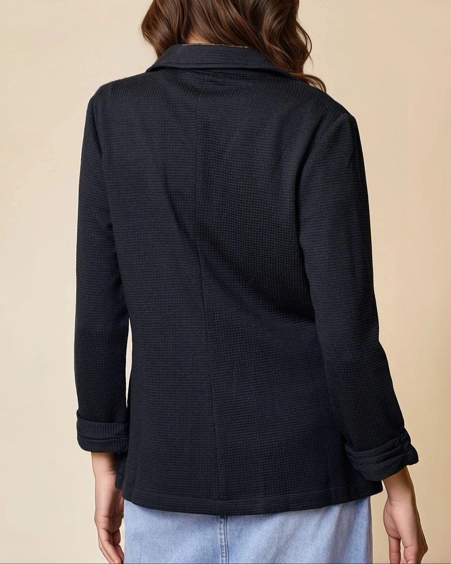 Woman wearing a black textured blazer over a white top and jean skirt on a beige background.