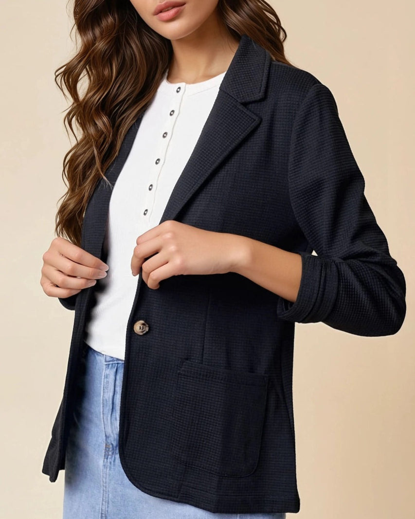 Woman wearing a black textured blazer over a white top and jean skirt on a beige background.