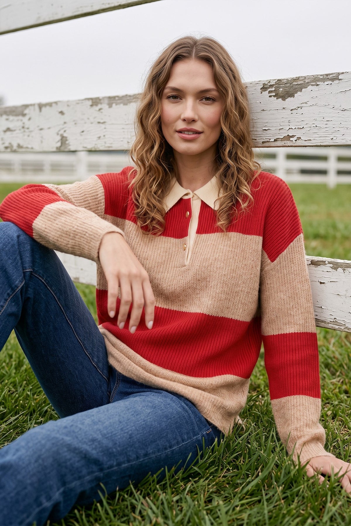 A woman sitting on green grass in front of a weathered white wooden fence, wearing a long-sleeve ribbed rugby-style sweater with horizontal stripes in red and beige, paired with blue jeans.