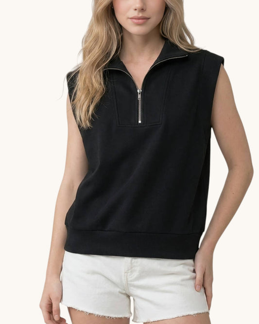 A woman wearing a sleeveless black half-zip pullover top with a mock neck and silver zipper, paired with white denim shorts, against a plain off-white background.