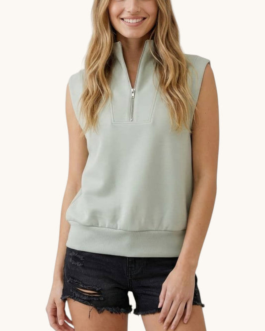 A woman wearing a sage green sleeveless quarter-zip sweatshirt and distressed black denim shorts, shown from the waist up against a light off-white background.