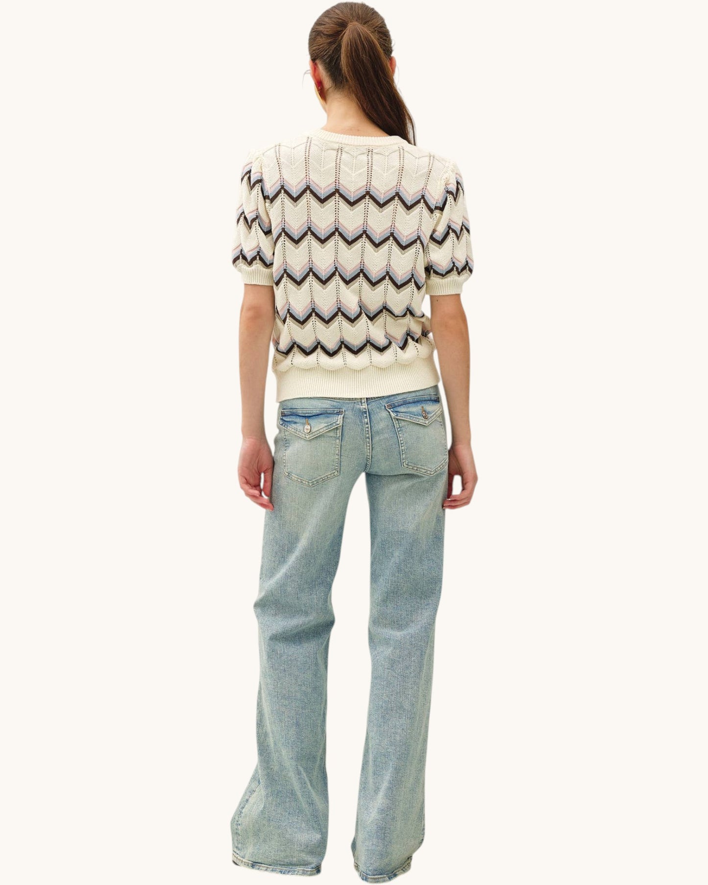 A woman shown from the back wearing a cream short-sleeve knit sweater with a multi-color zigzag pattern and light-wash blue jeans, against a plain off-white background.