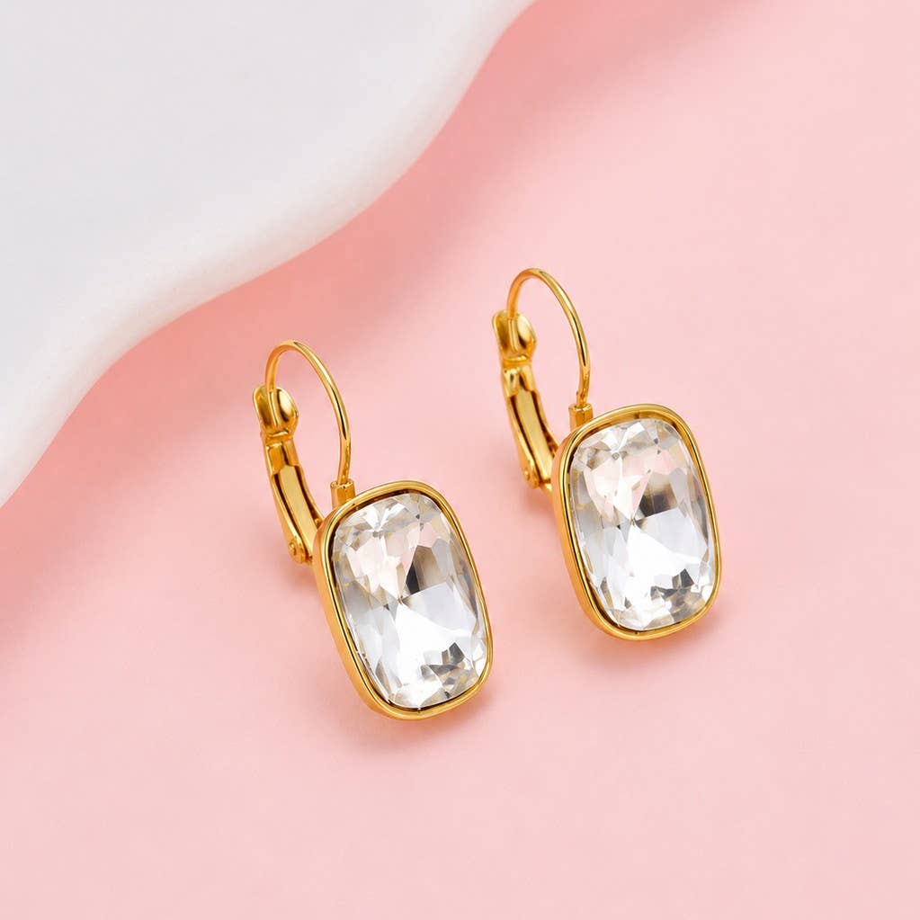 A pair of gold leverback earrings with large, clear, rectangular faceted crystals, displayed on a split pink and white background.