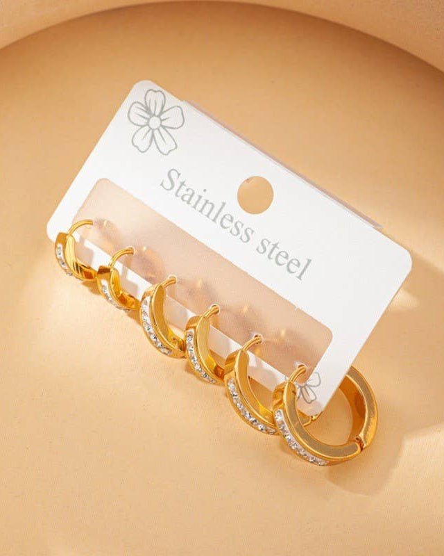 Set of gold hoop earrings with clear rhinestones on a white card labeled 'Stainless steel' against a beige background.