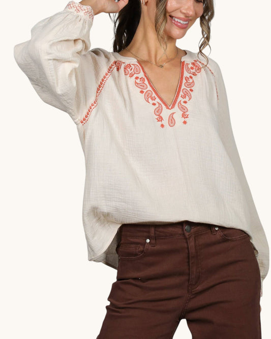 A woman wearing a loose off-white blouse with coral-red embroidered detailing around the V-neck and on the sleeve cuffs, paired with brown pants, against a plain light background.