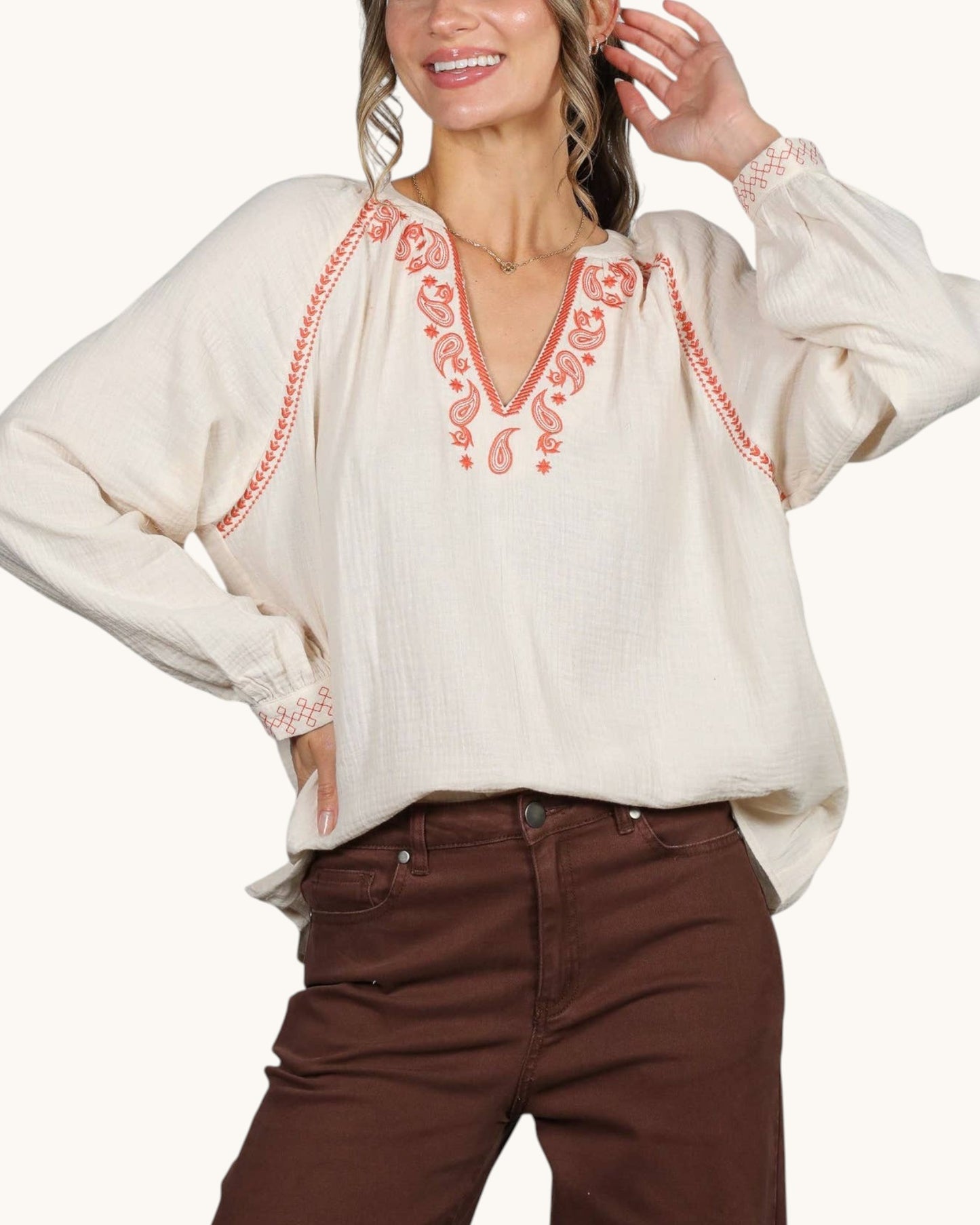 A woman wearing a cream-colored long-sleeve blouse with coral-red paisley embroidery around the V-neck and cuffs, paired with brown pants, against a plain light background.