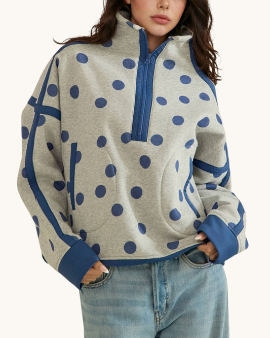 A woman wearing a light heather gray half-zip sweatshirt with navy blue polka dots and solid navy blue trim, paired with light-wash blue jeans against a plain off-white background.