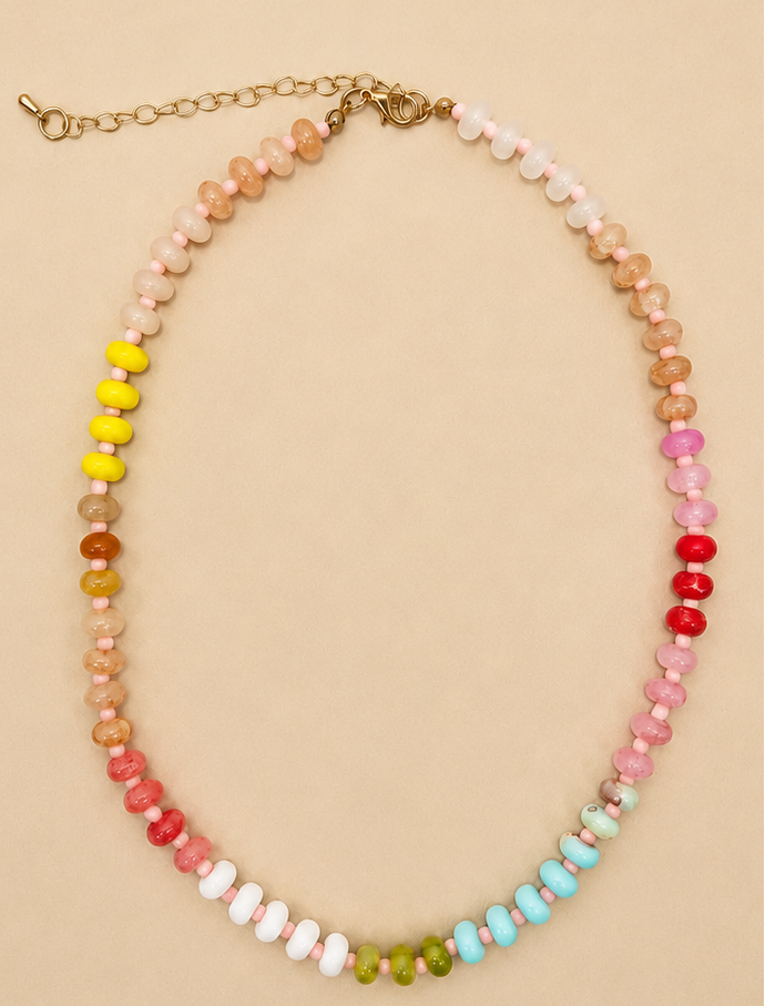 A beaded necklace with faceted round beads in a rainbow gradient, featuring a gold-tone chain clasp, displayed on a solid beige background.