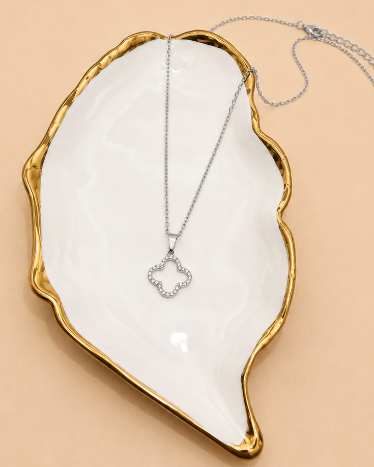 A silver-toned necklace with a small clover-shaped pendant set with clear stones, displayed on a white ceramic dish with a gold rim against a warm beige background.