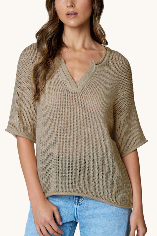 A woman wearing a beige open-knit short-sleeve V-neck sweater top with light blue jeans, shown from the waist up against a plain light background.