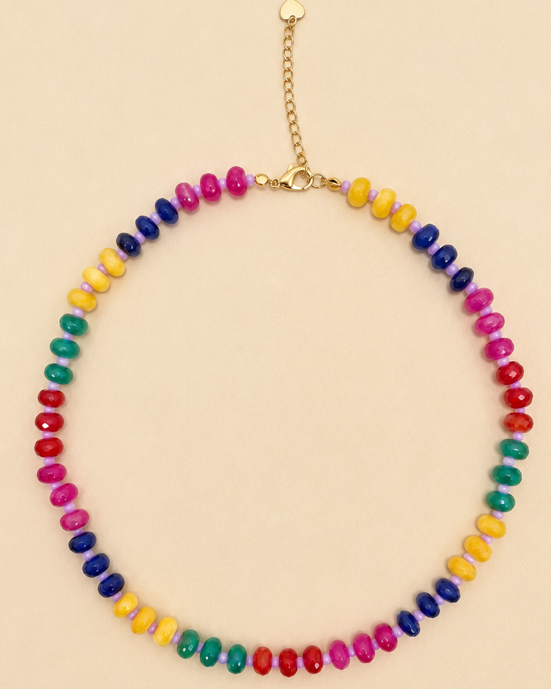 A beaded necklace with colorful faceted beads in a vibrant rainbow sequence, finished with a gold-tone clasp and extender chain, displayed on a solid beige background.