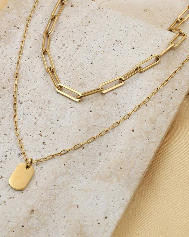 Gold chain necklace with a mini tag pendant on a textured beige stone surface.