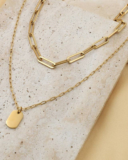 Gold chain necklace with a mini tag pendant on a textured beige stone surface.